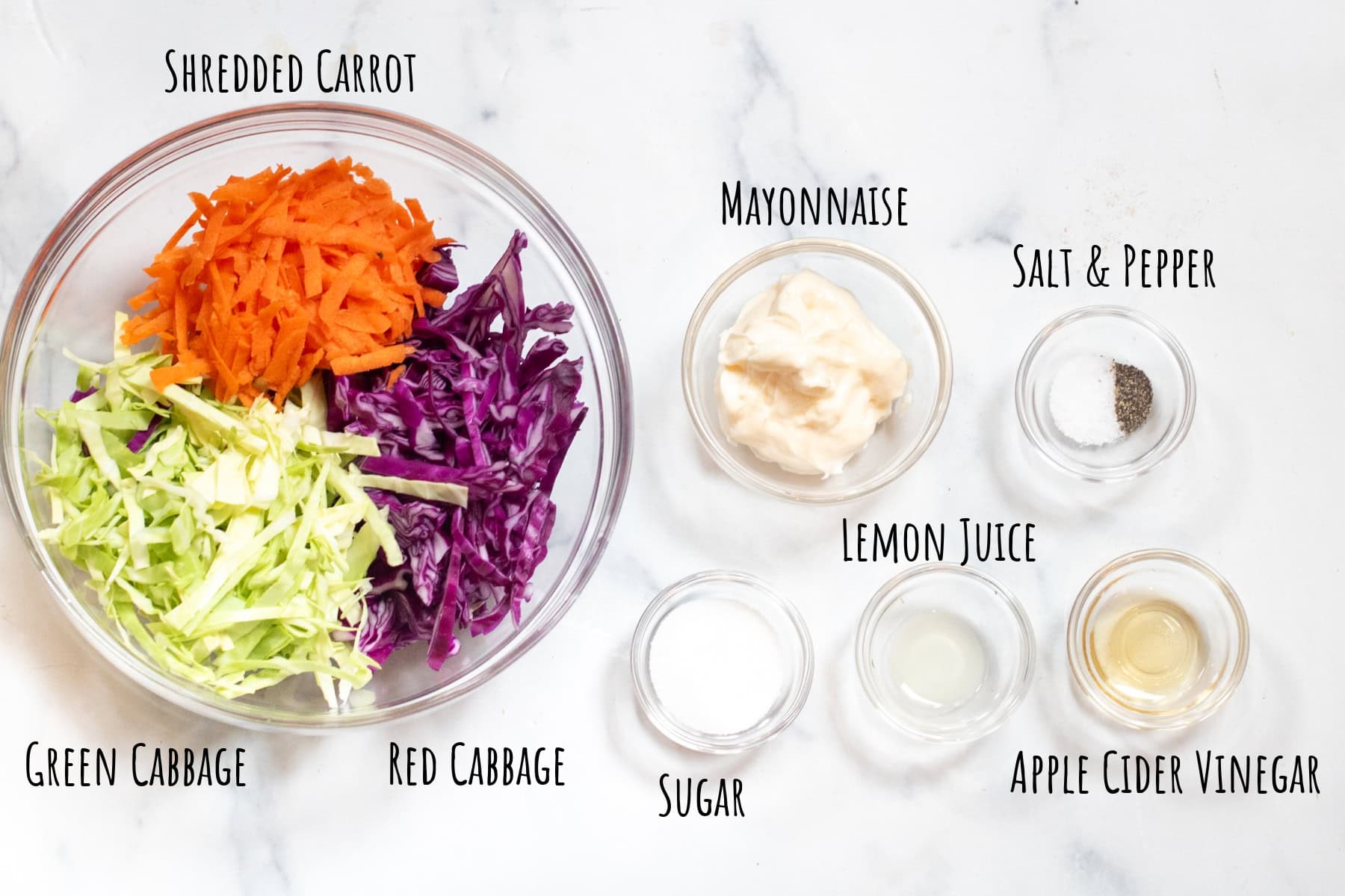 a bowl of green and purple cabbage with shredded carrots, mayo, salt and pepper, lemon juice, vinegar, and sugar all labeled.