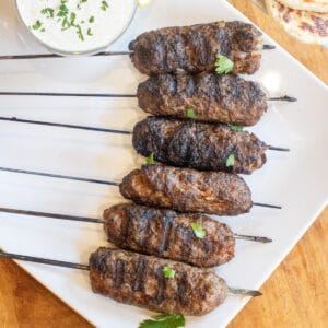 an overhead of grilled lamb kebabs on a plate with a bowl of yogurt sauce and next to some naan.