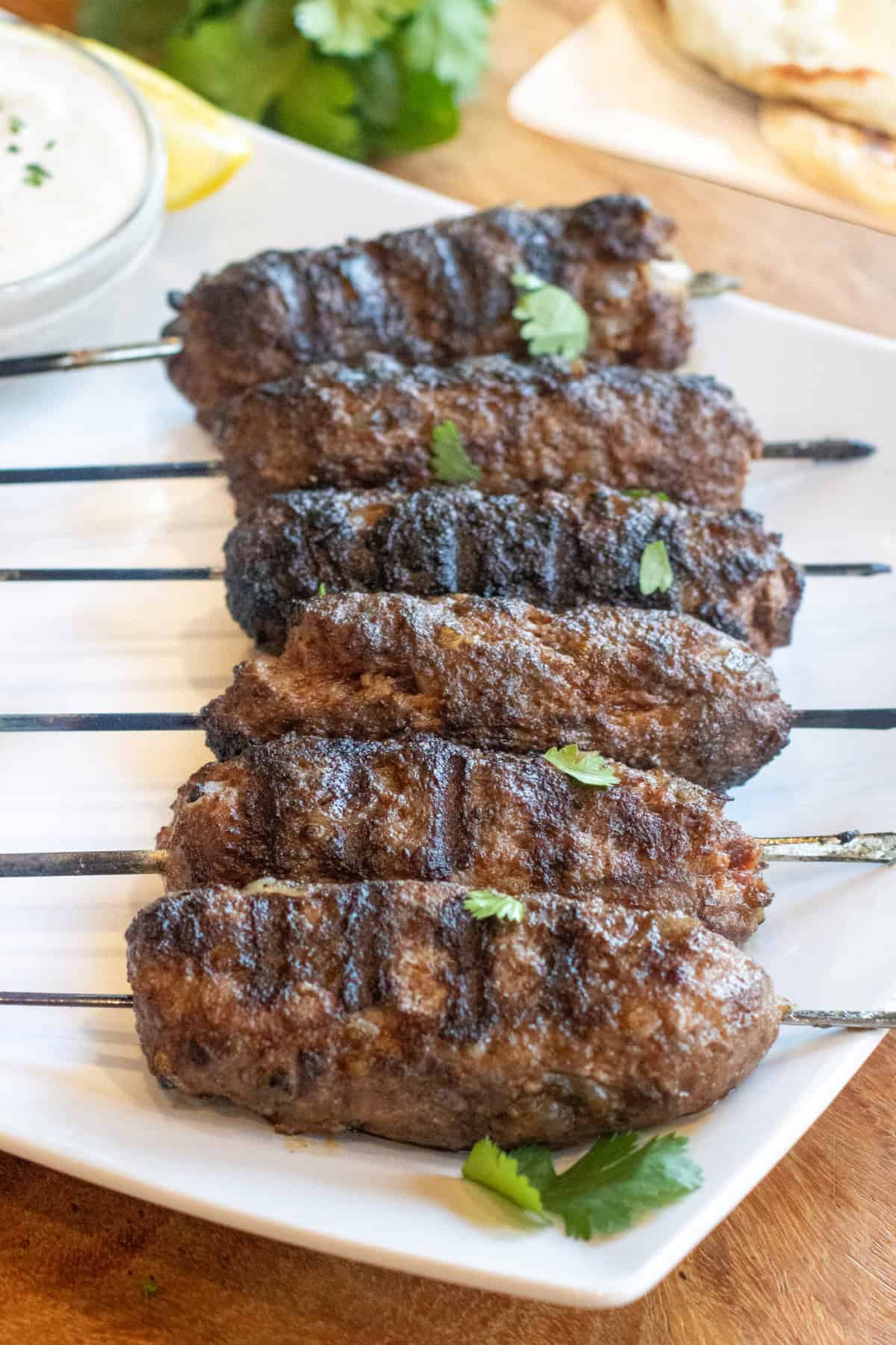 grilled cooked kofta skewers on a plate with a bowl of yogurt sauce and next to a plate of naan and some cilantro.