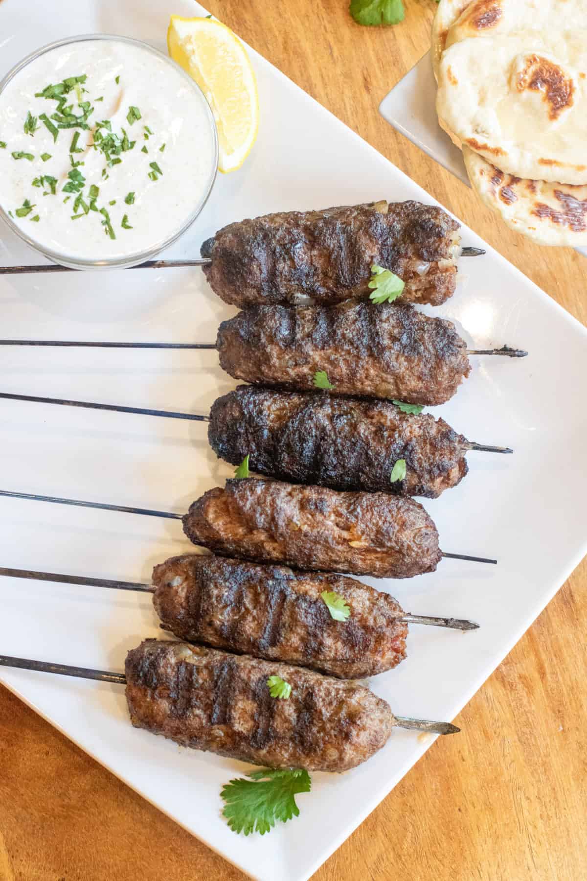 an overhead of grilled lamb kebabs on a plate with a bowl of yogurt sauce and next to some naan.