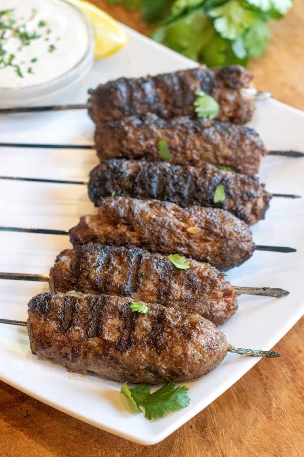 grilled cooked kofta skewers on a plate with a bowl of yogurt sauce and next to a plate of naan and some cilantro.