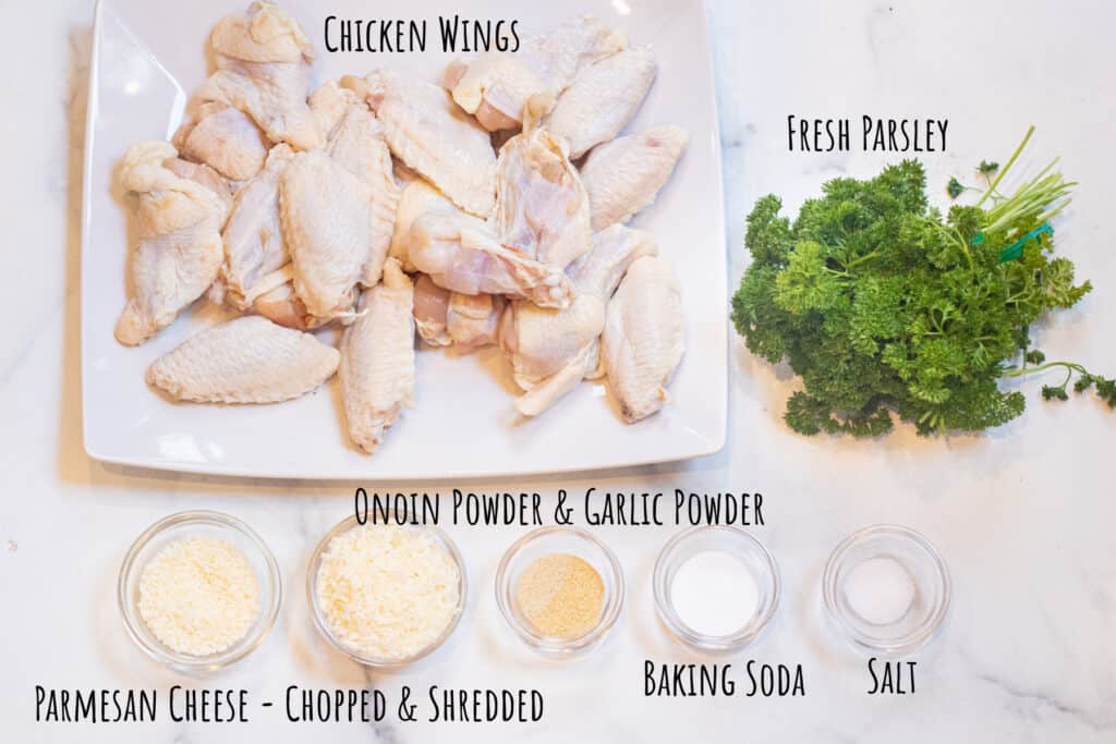 a plate of raw chicken wings, fresh parsley, spices and shredded parmesan cheese in bowls labeled.