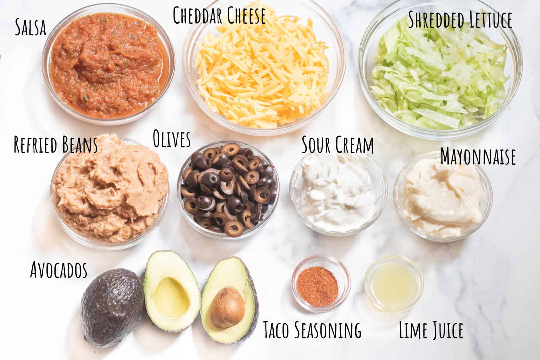 bowls of salsa, shredded cheese, lettuce, refried beans, olives, mayo, sour cream, taco seasoning, lemon juice, and halved avocados labeled.