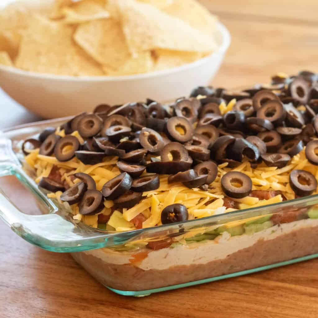 a clear baking dish with 7 layer taco dip in front of a bowl of chips.