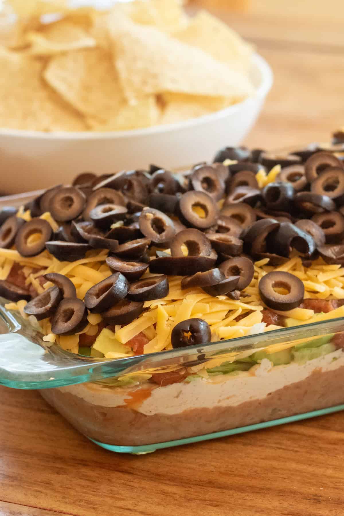 a clear baking dish with 7 layer taco dip in front of a bowl of chips.