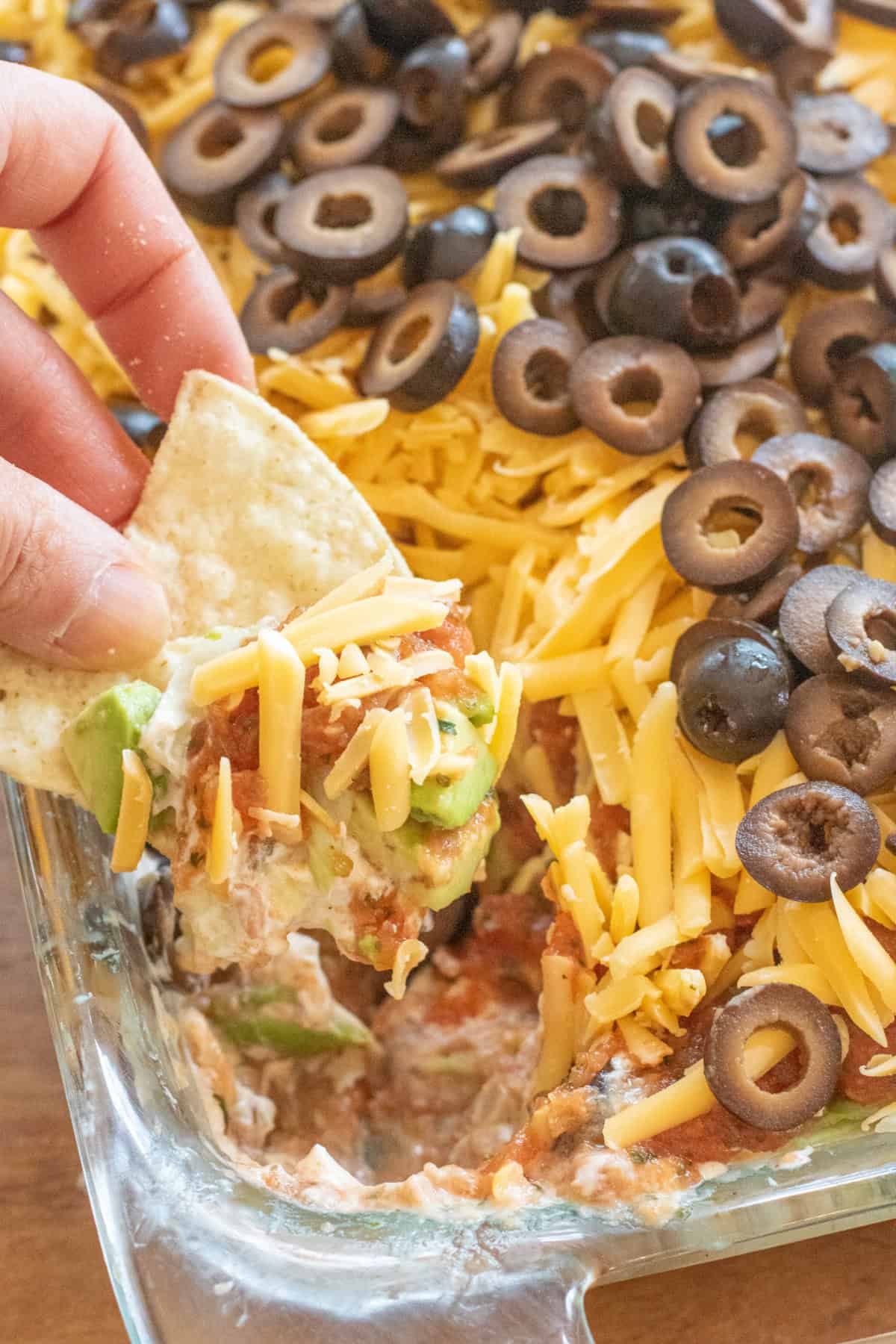a hand with a chip and a scoop of 7 layer dip from a tray of a layered dip.
