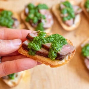 a hand holding a crostini with steak and chimichurri.