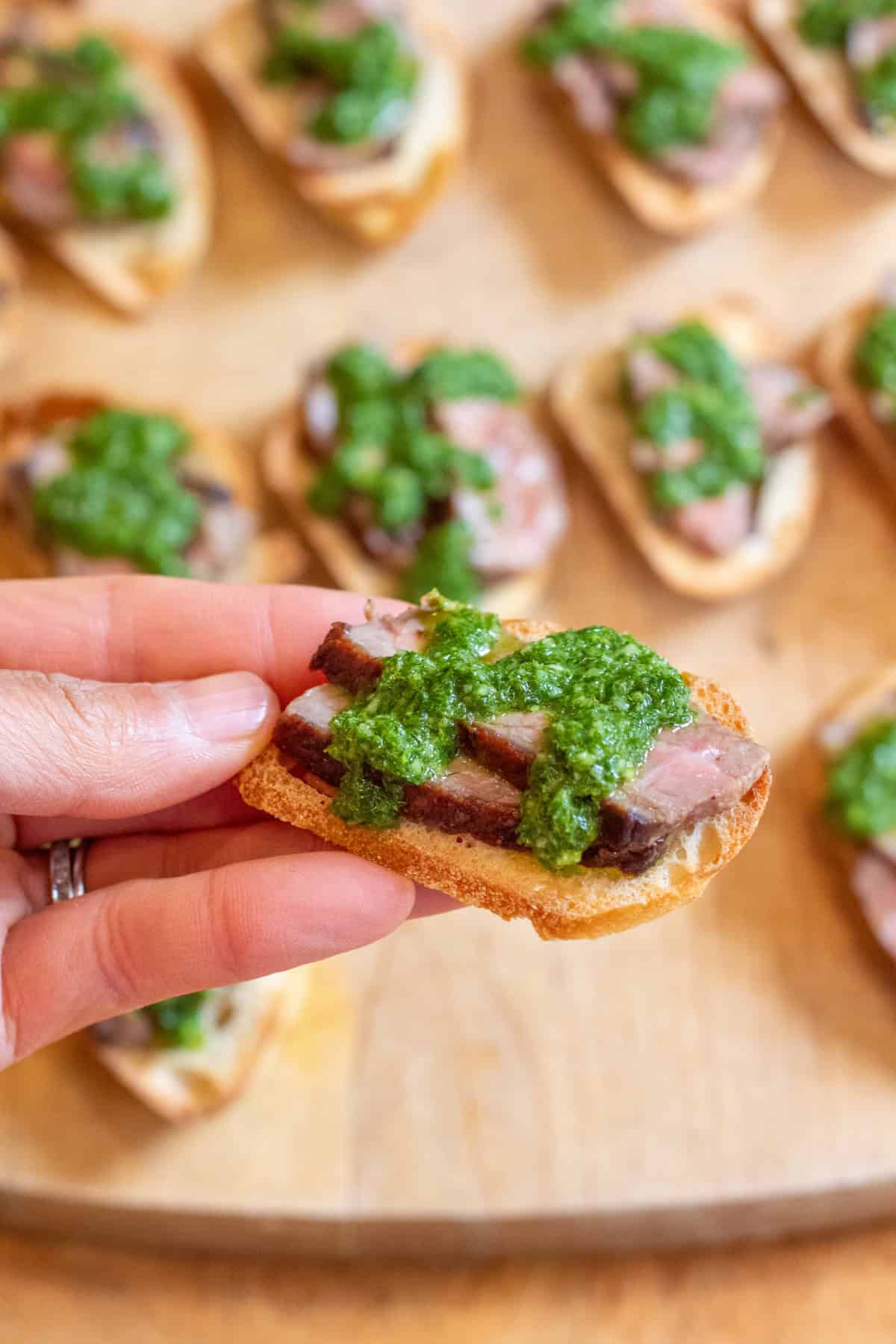 a hand holding a crostini with steak and chimichurri.