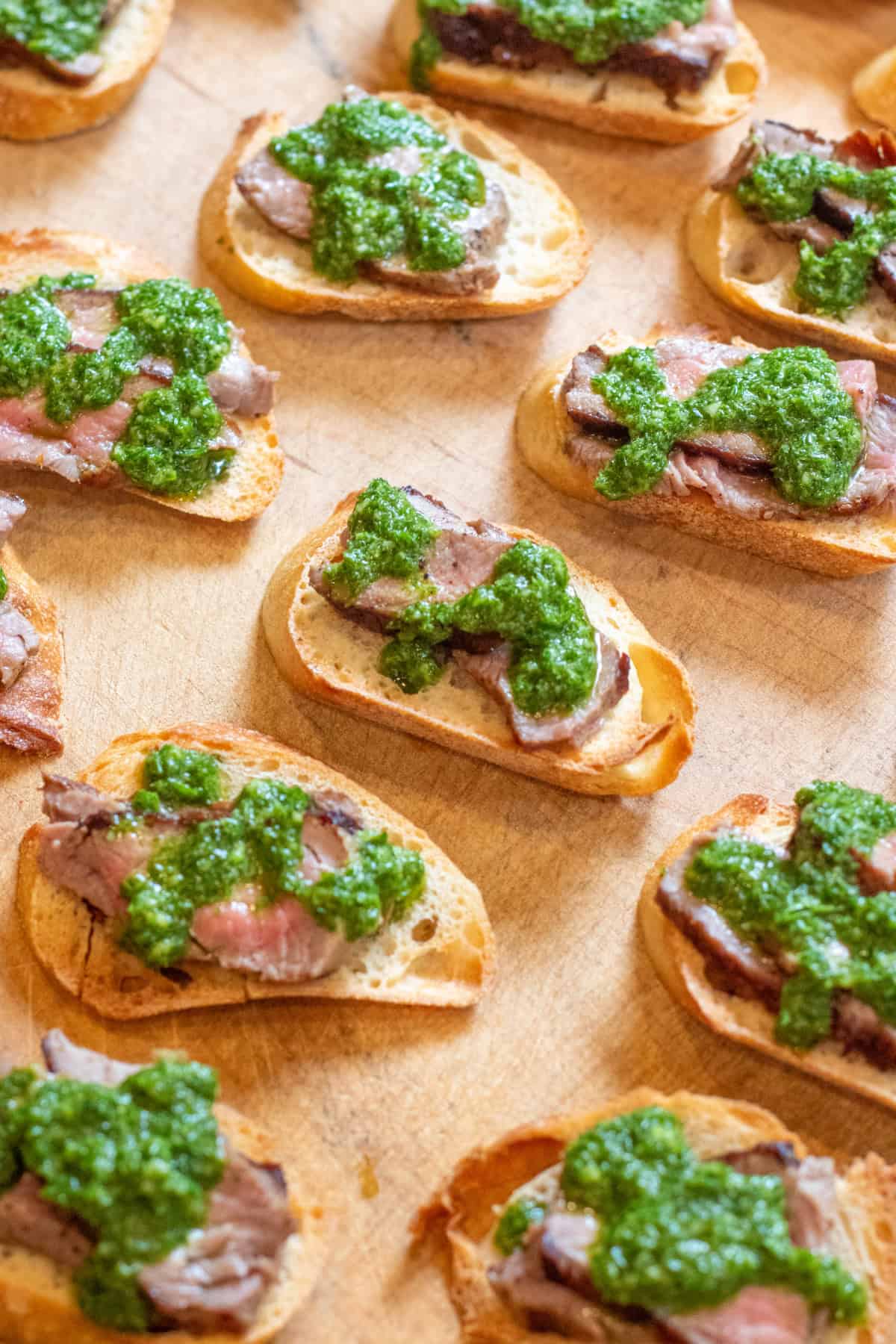 an overhead of a cutting board of chimichurri topped steak crostini.