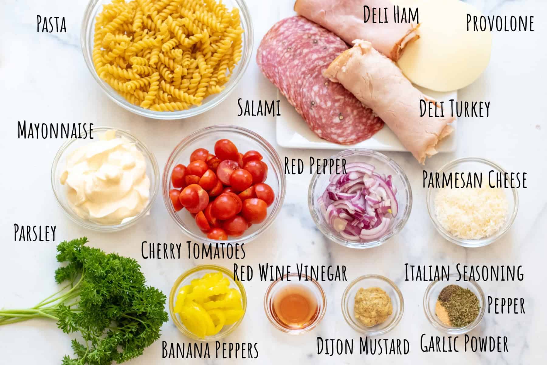 bowls of pasta, cherry tomatoes, red onion slices, mayo, shredded parm cheese, a plate of deli meats and provolone, banana peppers, vinegar, herbs, and fresh parsley, labeled.