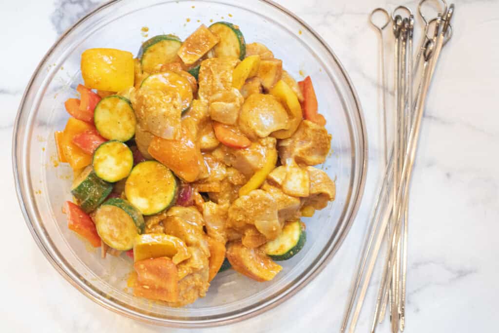 a bowl of marinaded chicken and veggies and metal skewers next to it.
