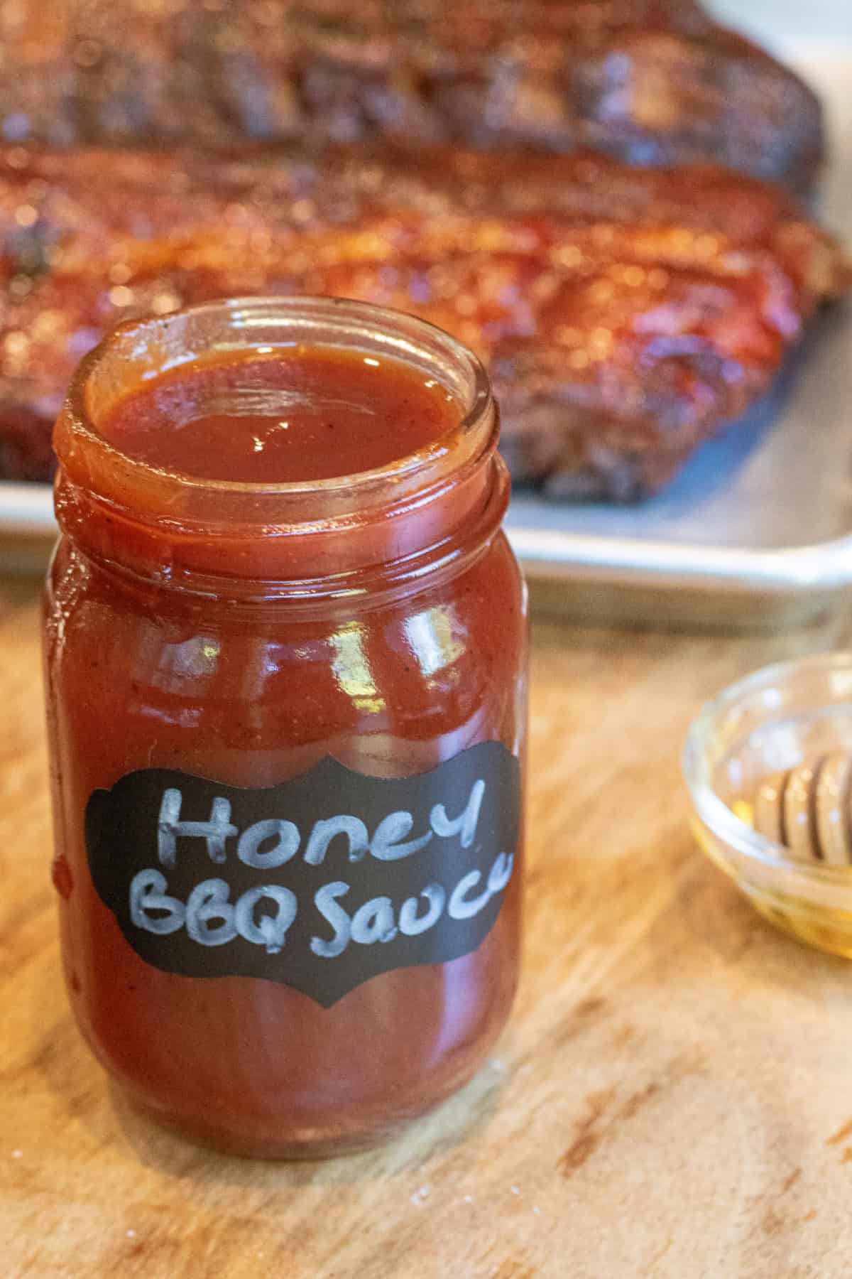 an open jar of honey bbq sauce in front of a tray of ribs.