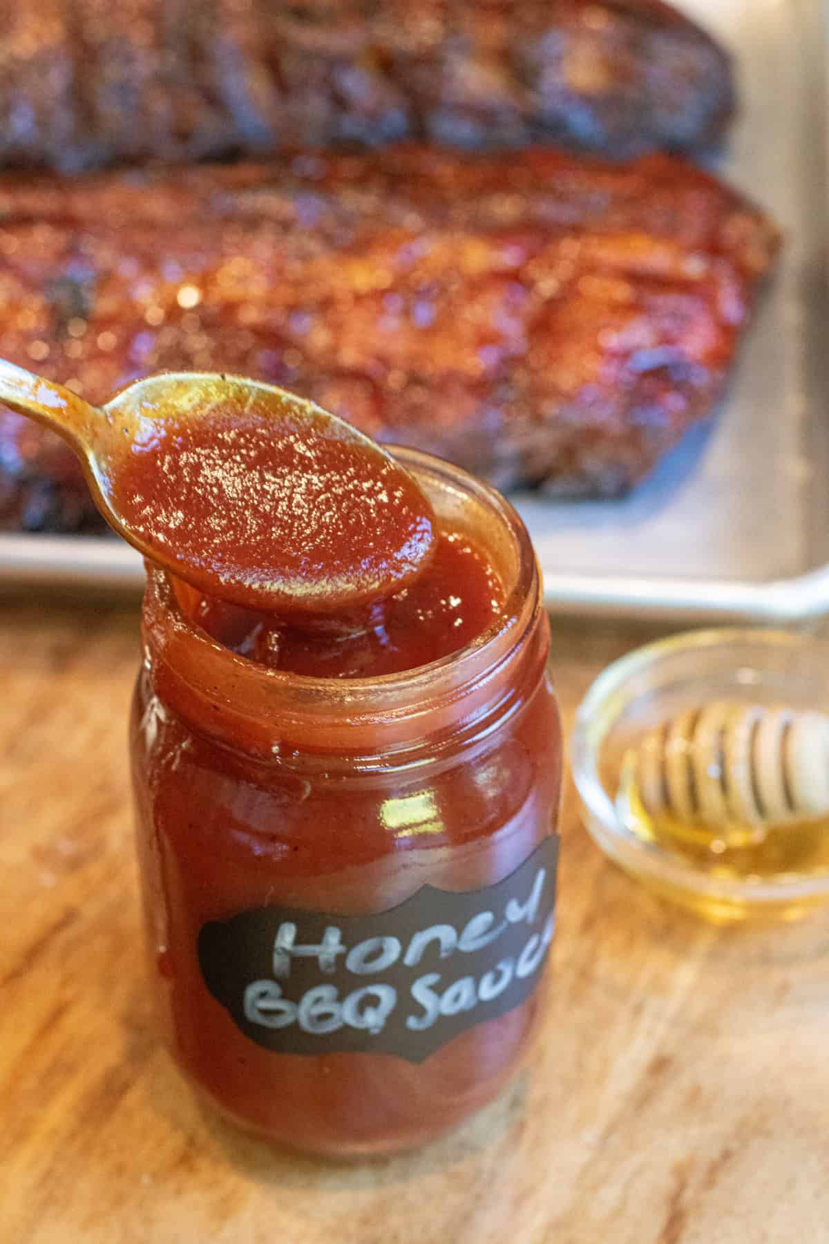 an open jar of honey bbq sauce with a spoon in front of a tray of ribs.