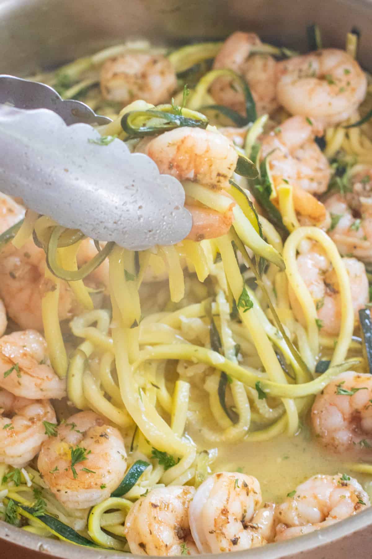 an overhead of a pan with cooked shrimp and zoodles and tongs holding some up.