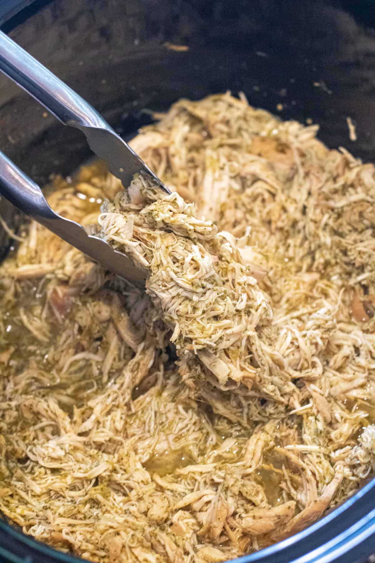 a crock pot with shredded chicken and tongs holding some up.