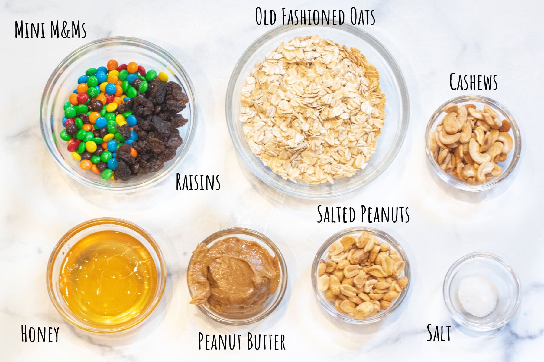 bowls of mini M&Ms, oats, honey, peanut butter, cashews, peanuts, and salt in bowls all labeled.