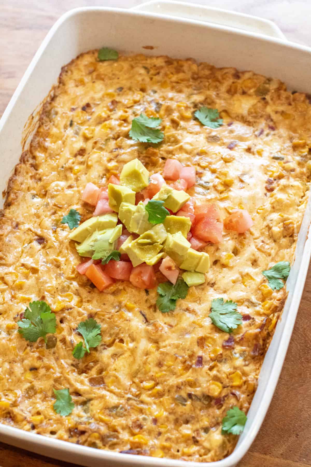 a baking dish of cheesy corn dip topped with diced tomatoes, avocado, and fresh cilantro.