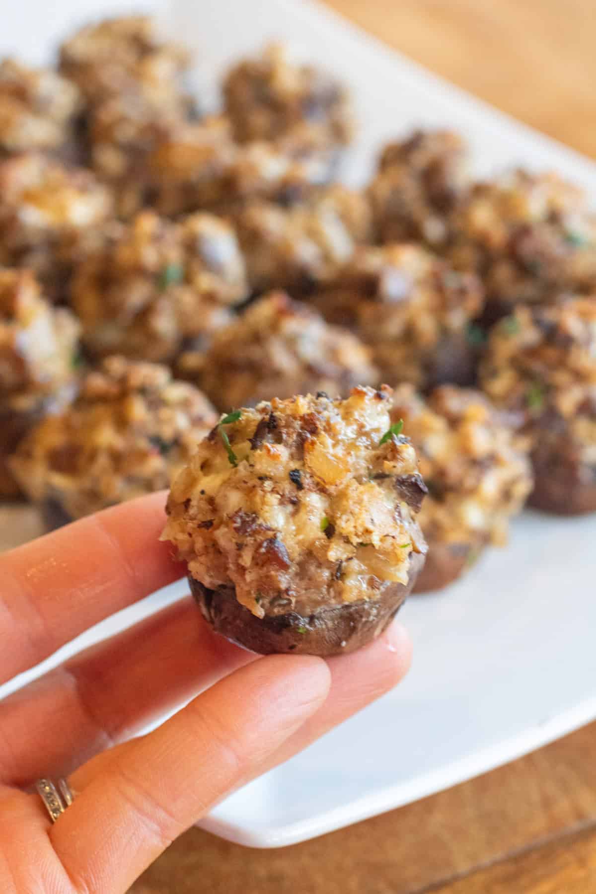 a hand holding a baked stuffed mushroom in front of a plate of baked stuffed mushrooms.