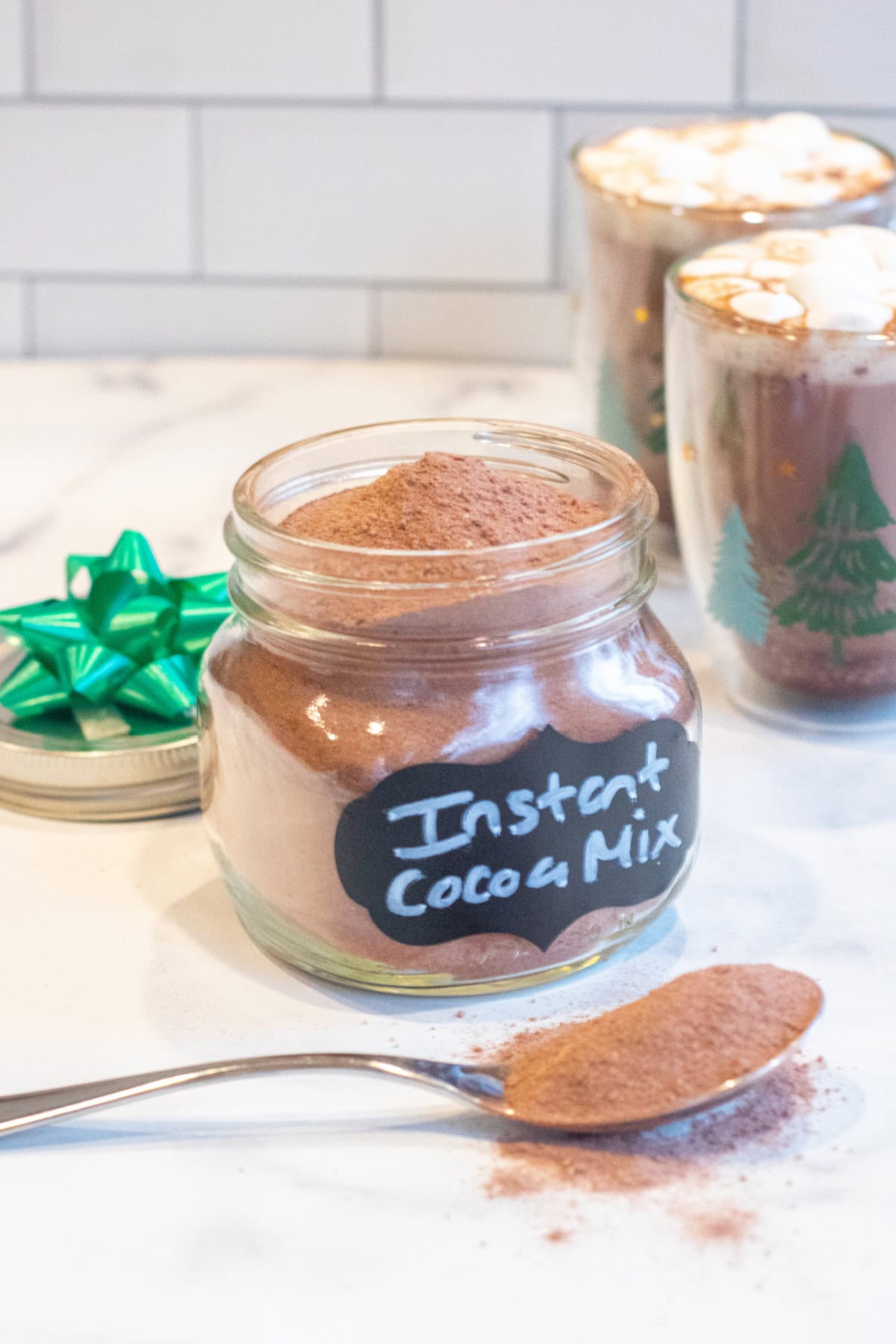 an open mason jar with a label of instant cocoa mix in front of a two mugs of cocoa with mini marshmallows and spoon in front of the jar.