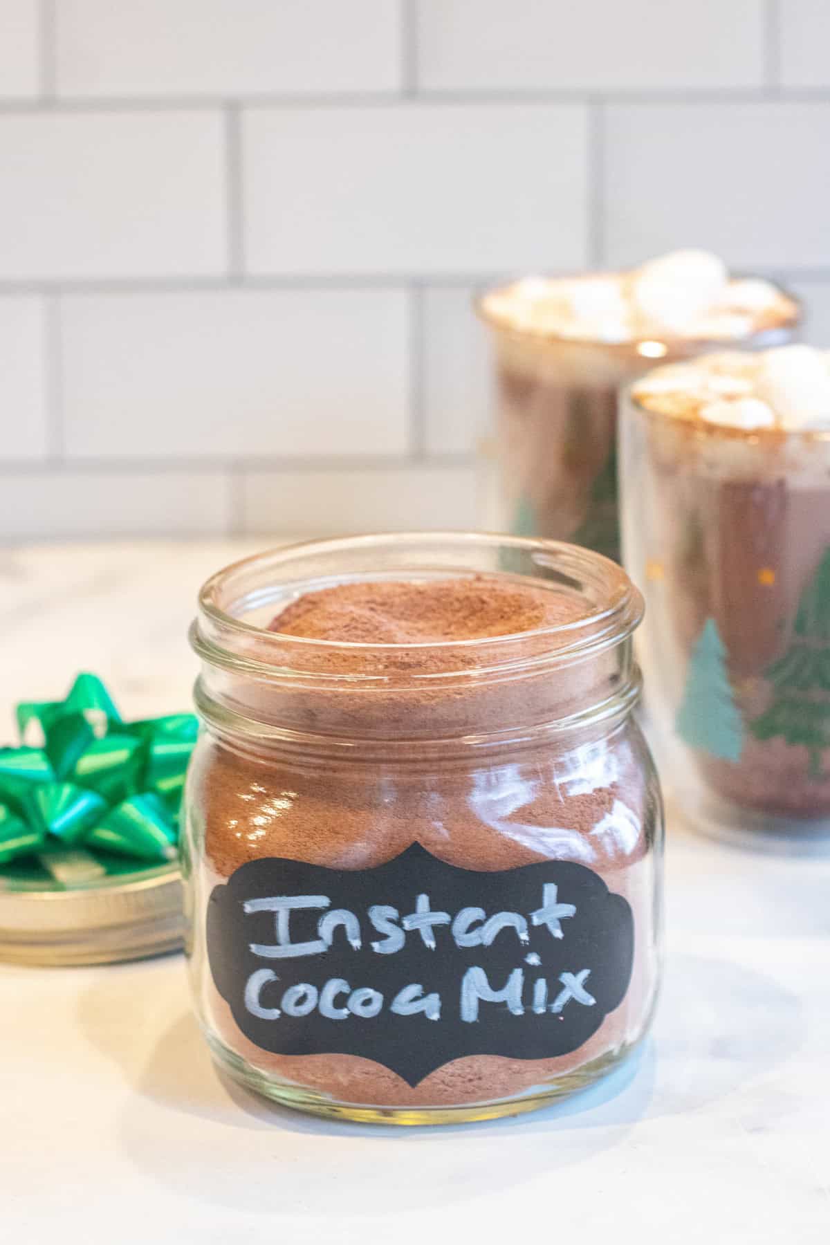 an open mason jar with a label of instant cocoa mix in front of a two mugs of cocoa with mini marshmallows.