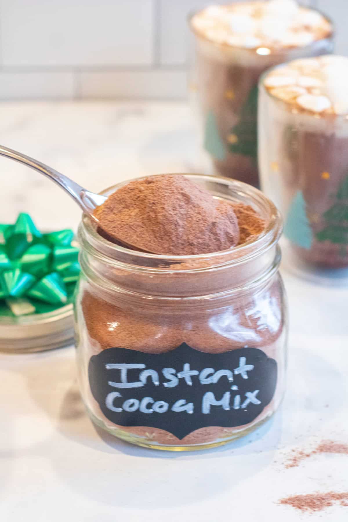 an open mason jar with a label of instant cocoa mix with a spoonful of it coming out in front of a two mugs of cocoa with mini marshmallows.