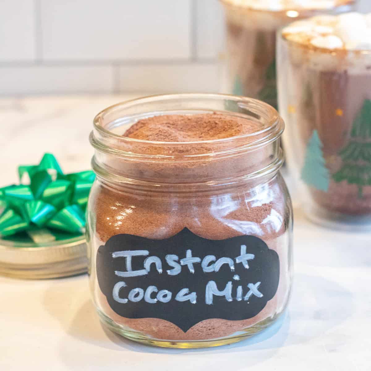 an open mason jar with a label of instant cocoa mix in front of a two mugs of cocoa with mini marshmallows.