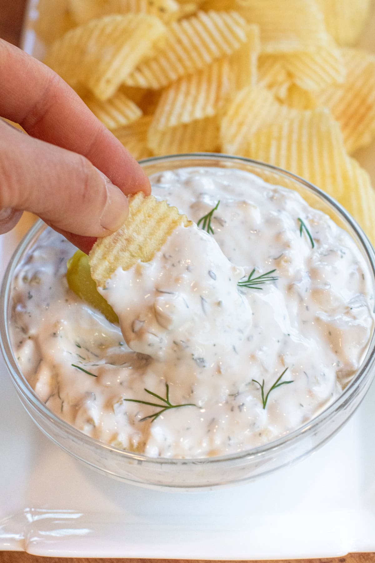 a bowl of creamy dip with a pickle and fresh dill on a plate of chips and a hand holding a chip with dip.
