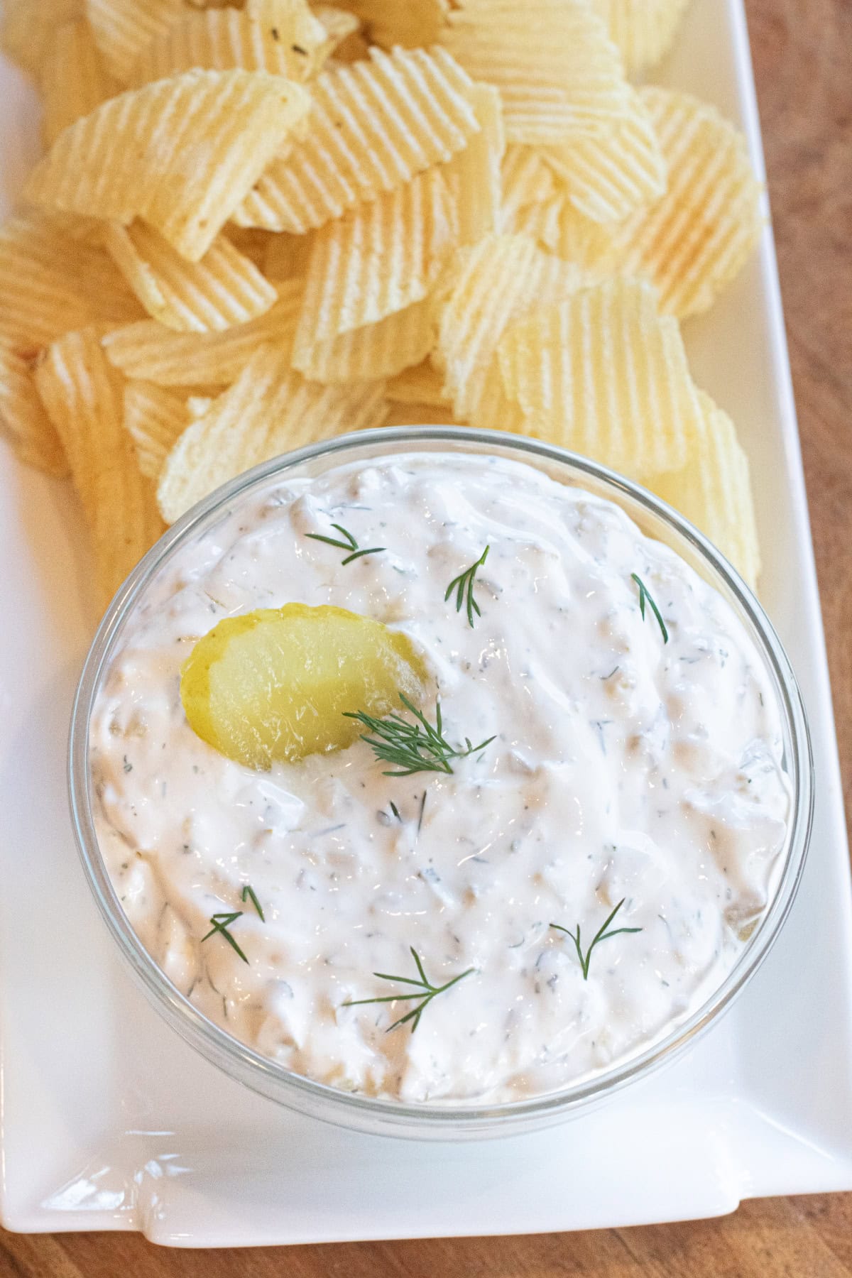 a bowl of creamy dip with a pickle and fresh dill on a plate of chips.