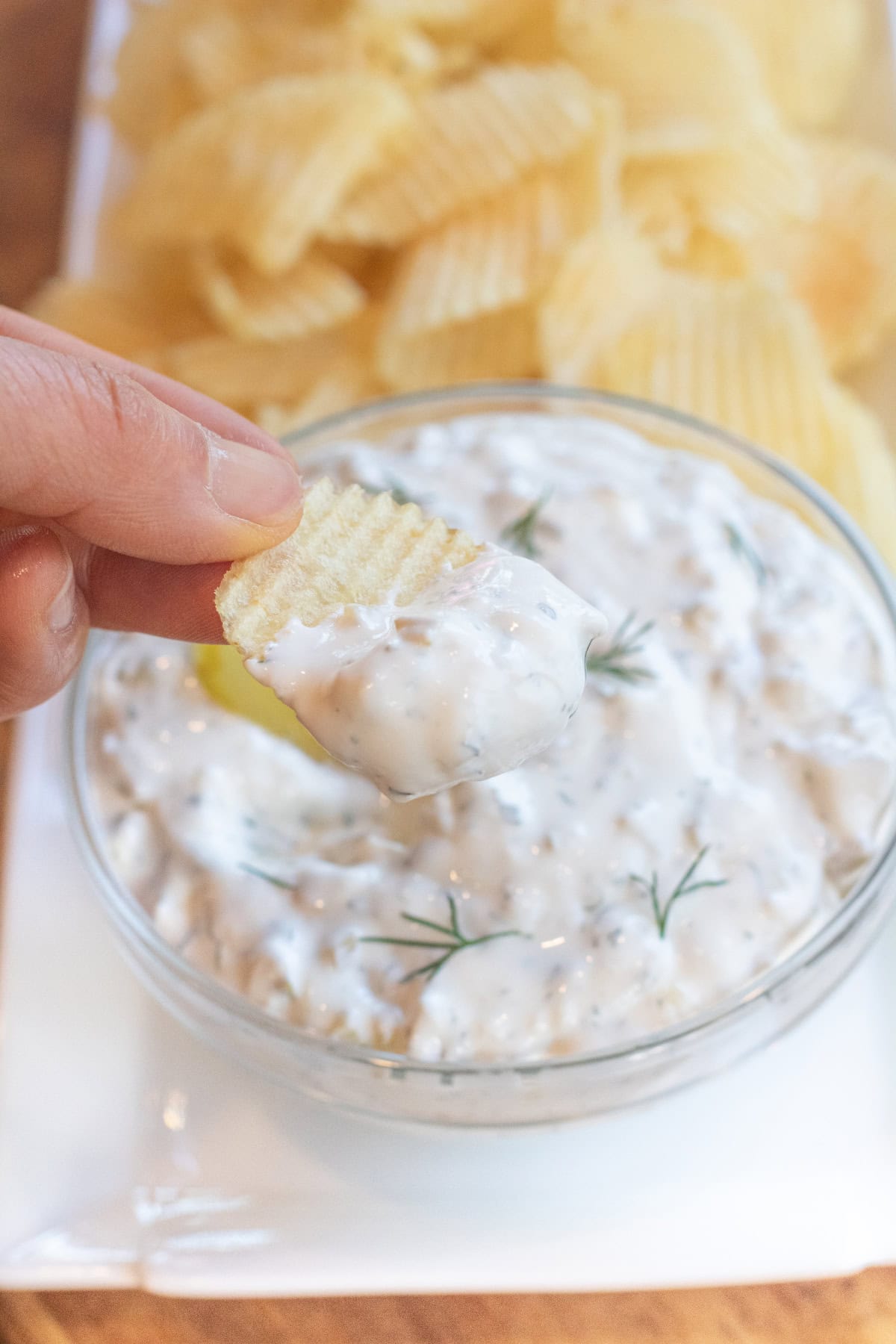 a bowl of creamy dip with a pickle and fresh dill on a plate of chips and a hand holding a chip with dip.