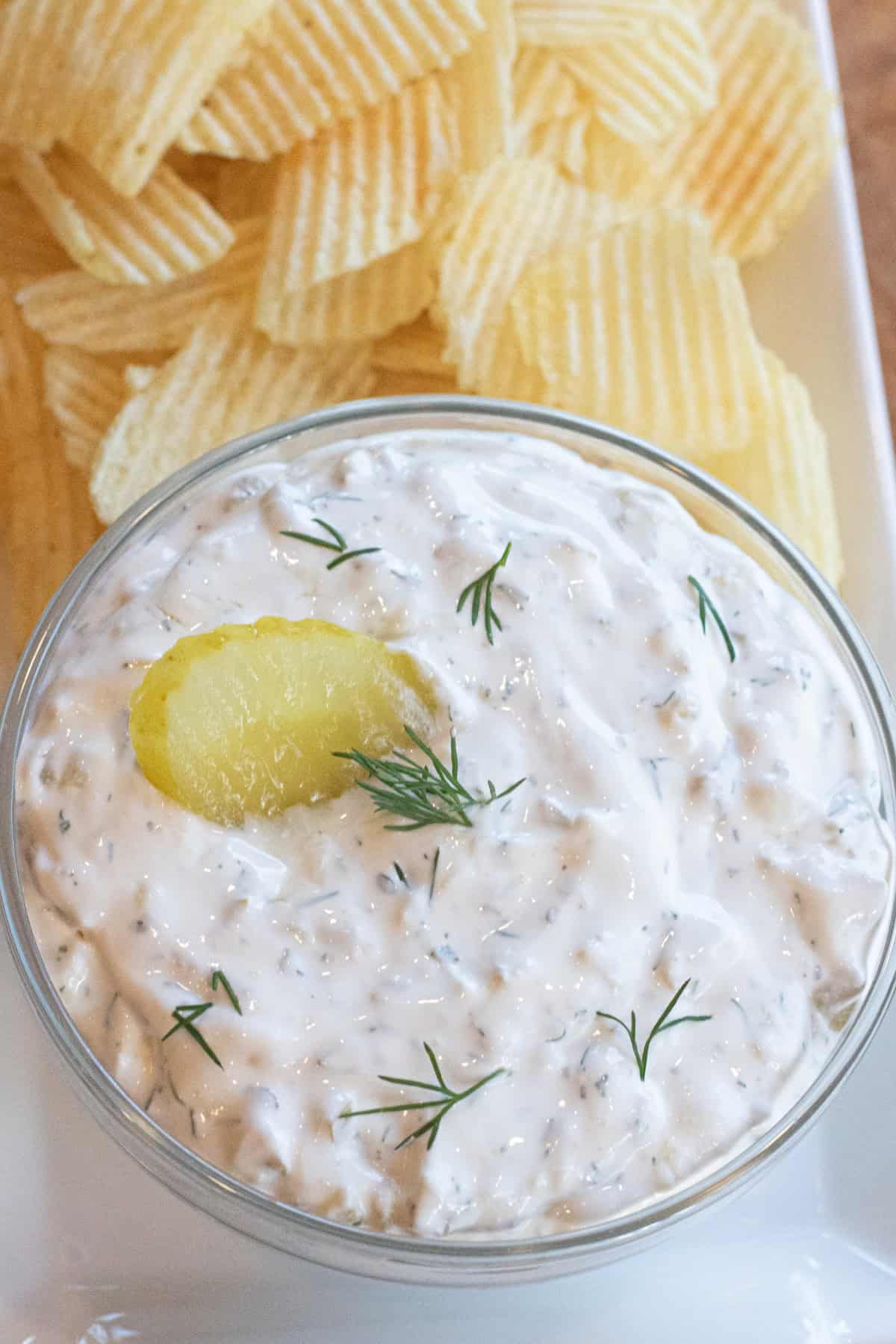 a bowl of creamy dip with a pickle and fresh dill on a plate of chips.