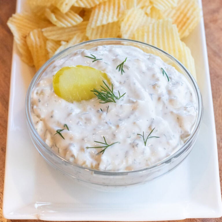 a bowl of creamy dip with a pickle and fresh dill on a plate of chips.