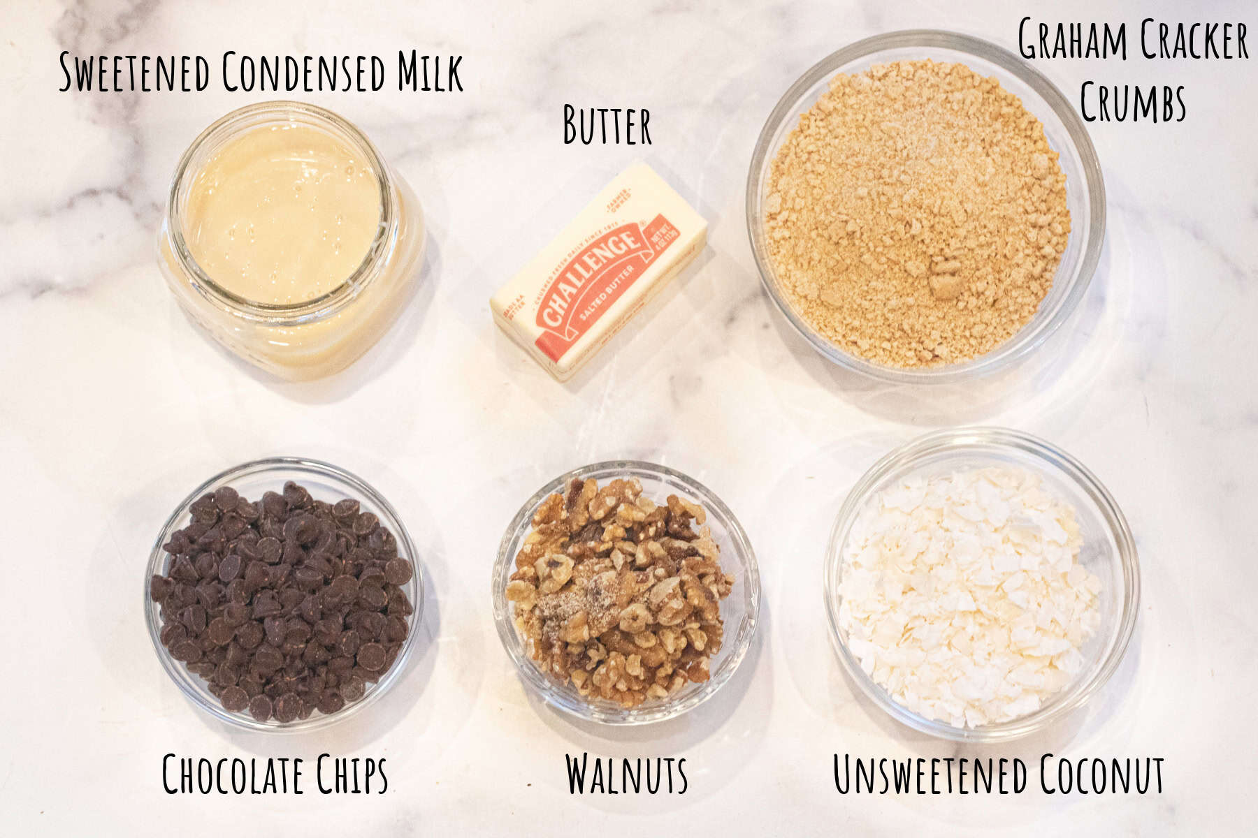bowls of graham cracker crumbs, chocolate chips, walnuts, coconut flakes, butter, and sweetened condensed milk all labeled.