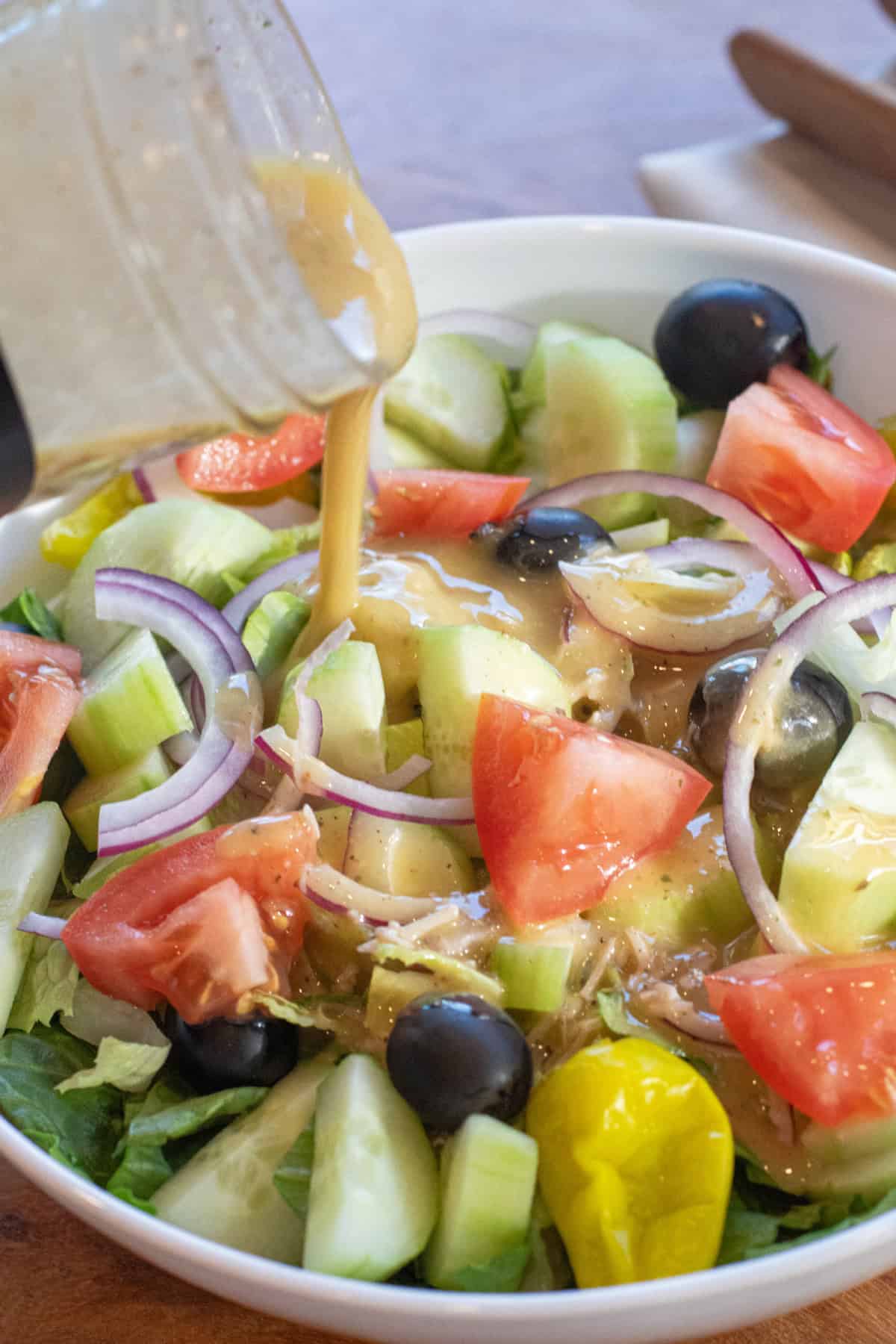 creamy Italian dressing being poured onto a big bowl of green salad with cucumbers, tomatoes, peppercinis, olives, and red onion.