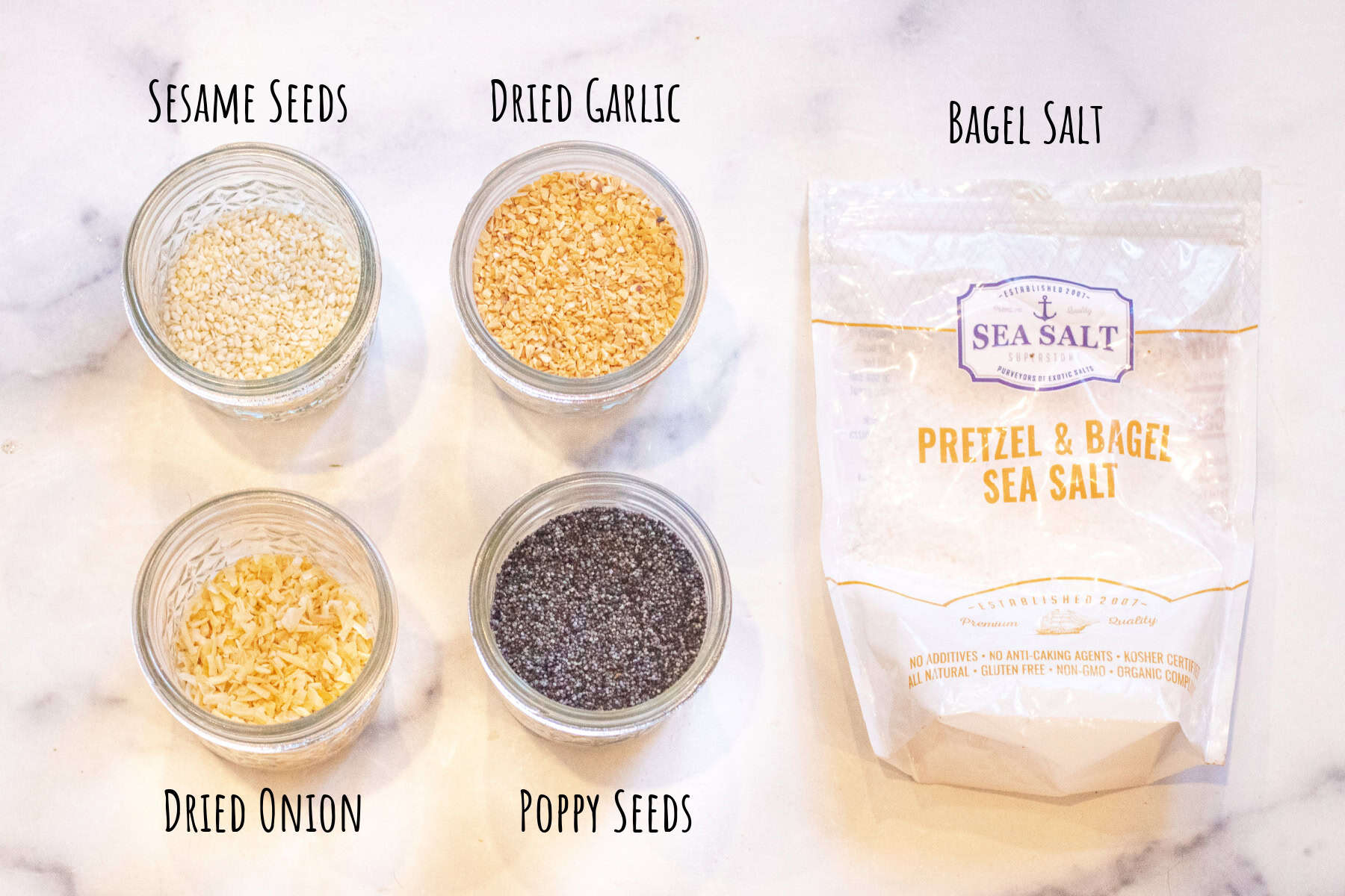 open jars of sesame seeds, dried garlic, onion, and poppy seeds and a bag of pretzel salt.
