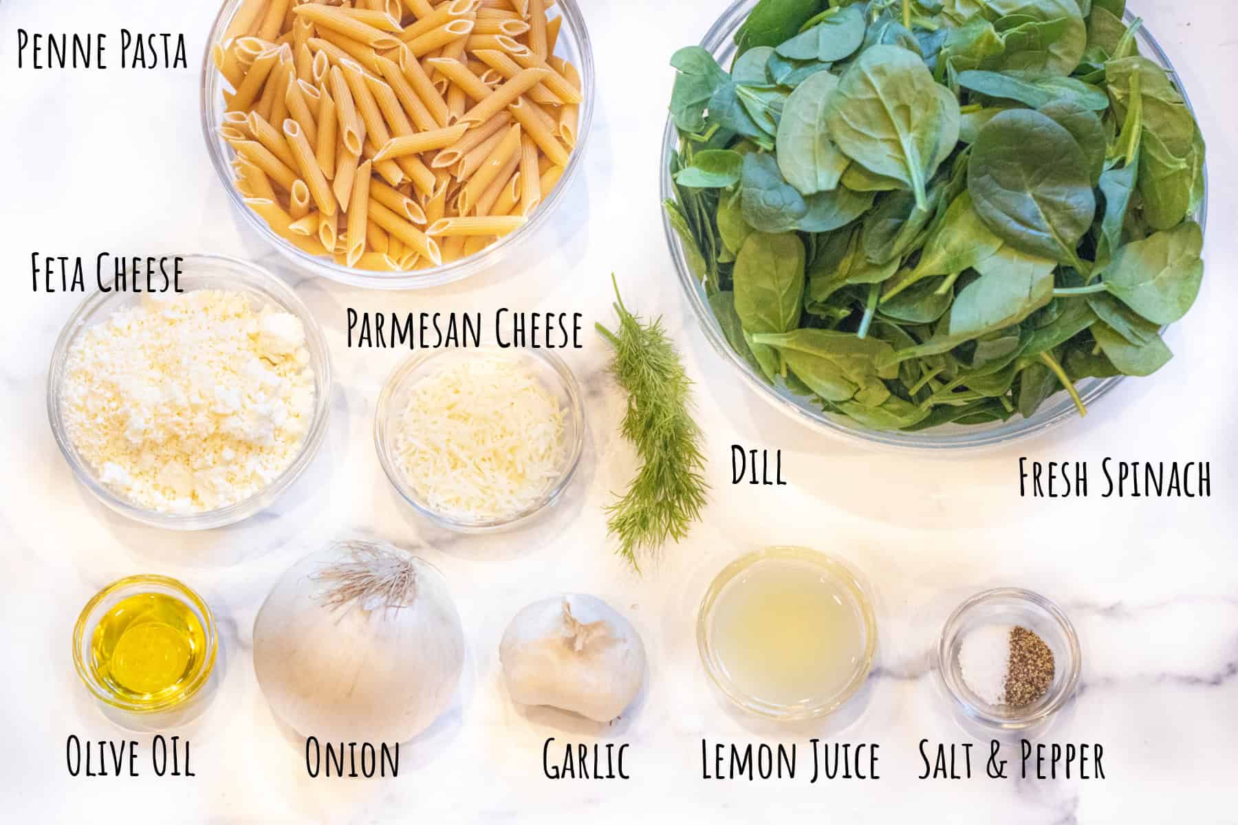 bowls of uncooked penne pasta, raw spinach, shredded parmesan cheese, feta, and onion, garlic, and bowls of salt and pepper, lemon juice, olive oil, fresh dill.
