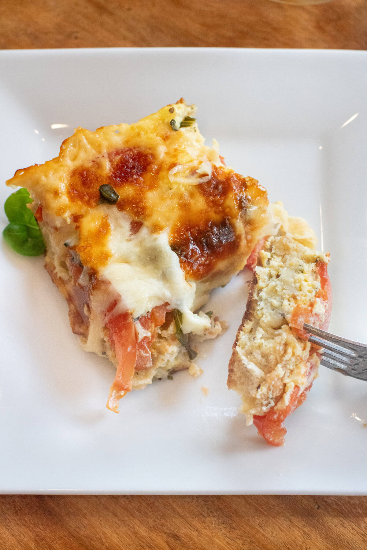 a piece of egg and tomato cheese and basil casserole on a plate in front of a baking dish of casserole with a fork and a piece of it.