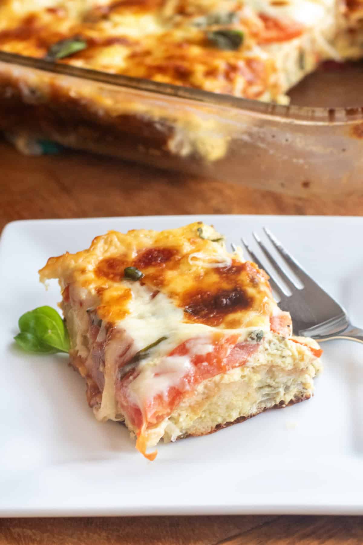 a piece of egg and tomato cheese and basil casserole on a plate in front of a baking dish of casserole.