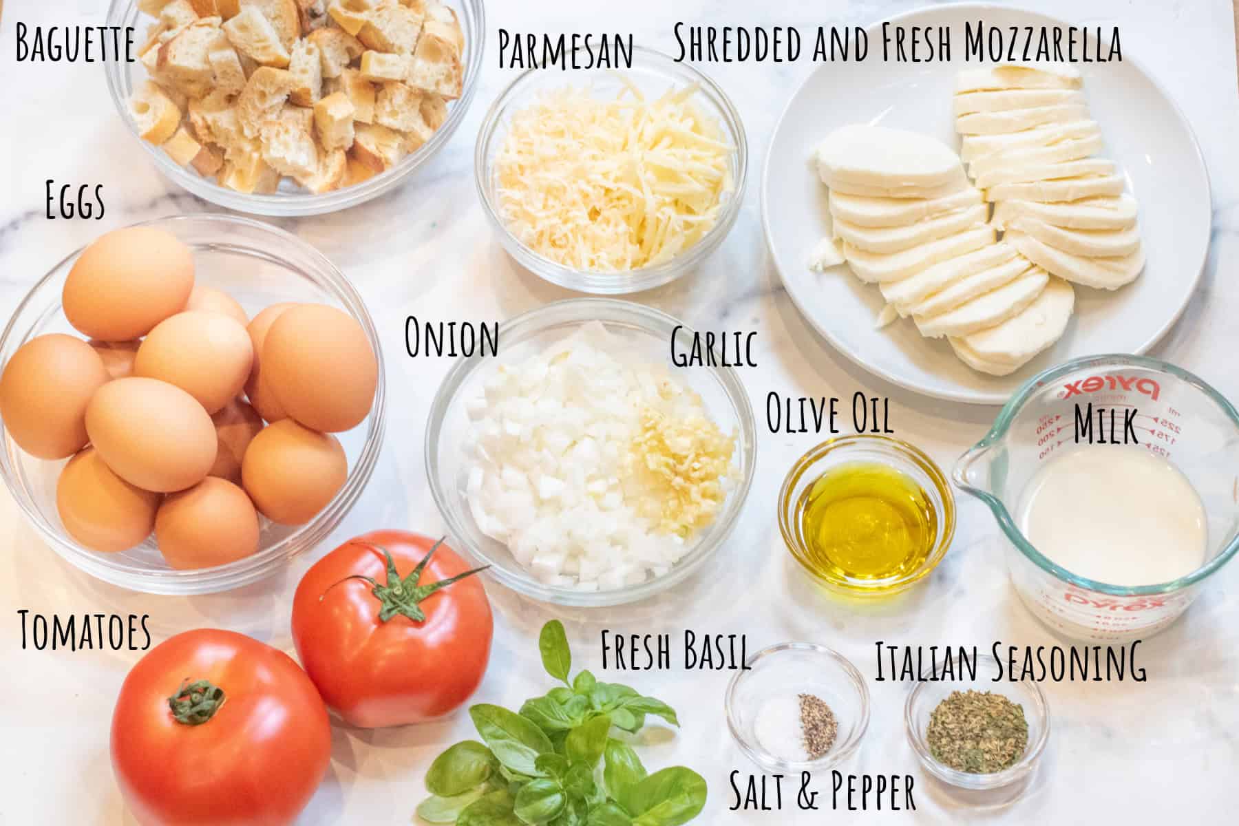 bowls of diced baguettes, eggs, cheeses, sliced fresh mozzarella, diced garlic and onion, seasoning, olive oil, salt and pepper, fresh basil, and two large tomatoes.