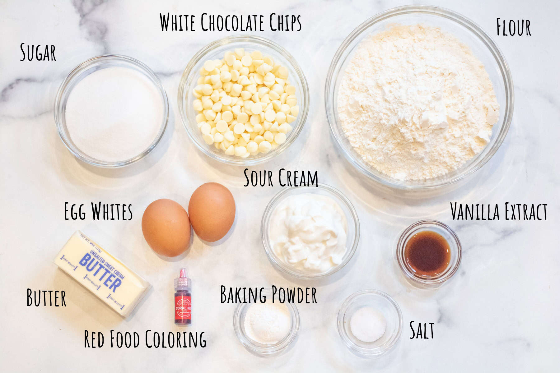 bowls of flour, white chocolate chips, sugar, eggs, vanilla extract, butter, baking powder, salt, and red food coloring labeled.