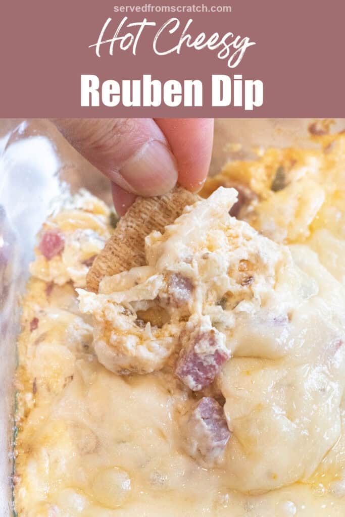 a hand holding a cracker dipping it into a Reuben dip with melted cheese and Pinterest pin text.