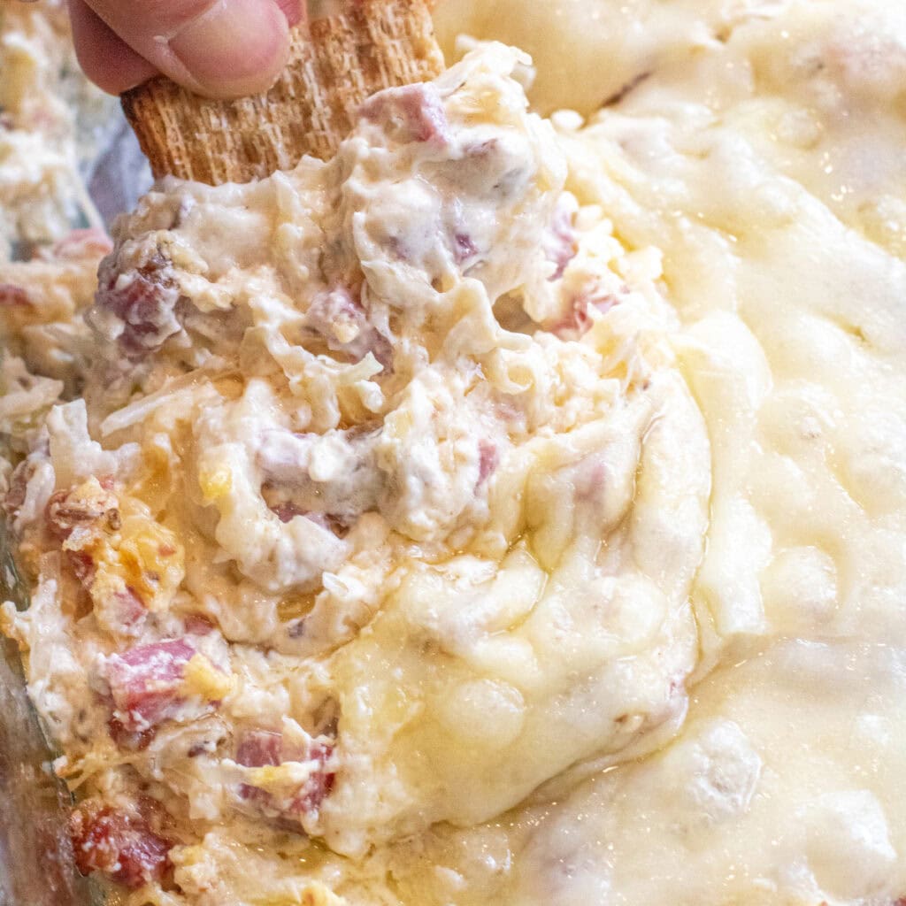 a hand holding a cracker dipping it into a Reuben dip with melted cheese.