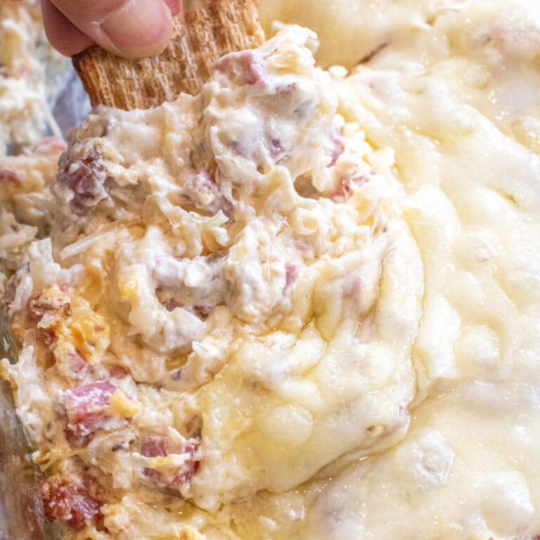a hand holding a cracker dipping it into a Reuben dip with melted cheese.