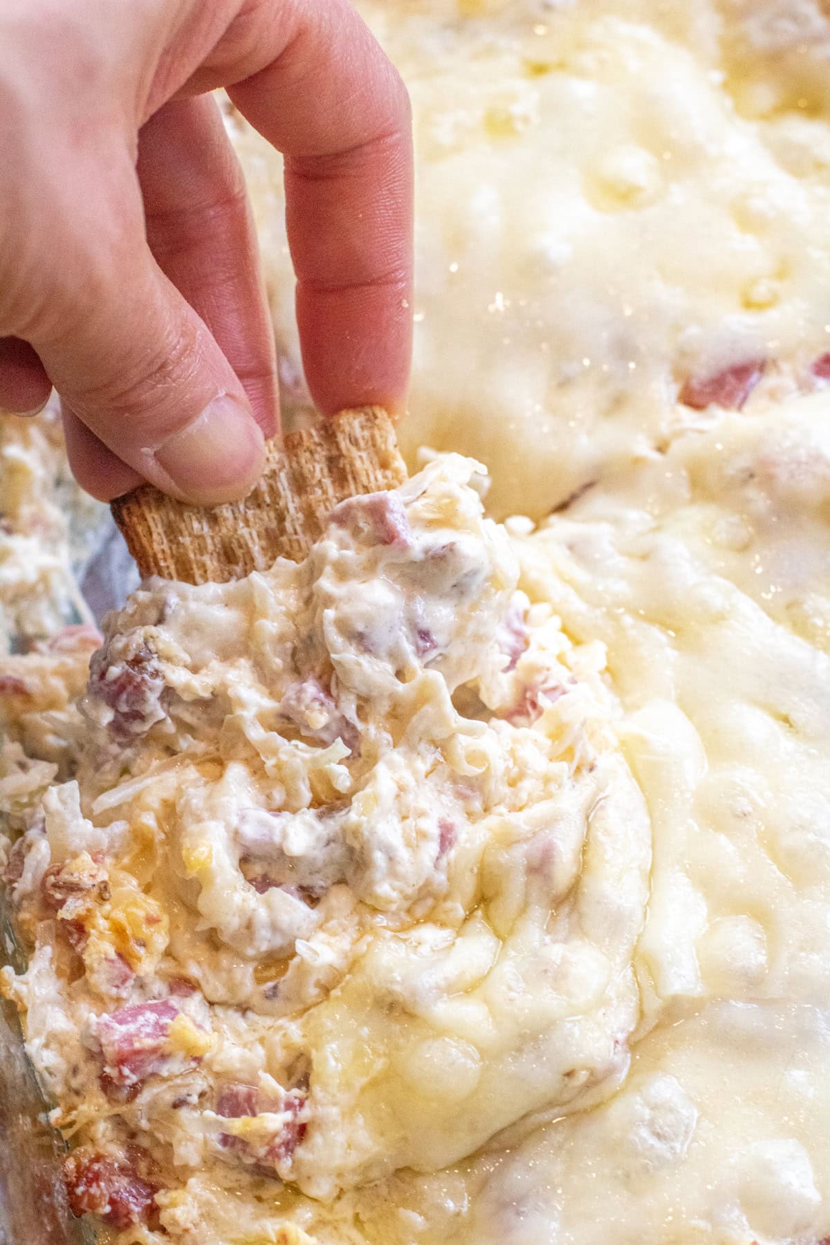 a hand holding a cracker dipping it into a Reuben dip with melted cheese.