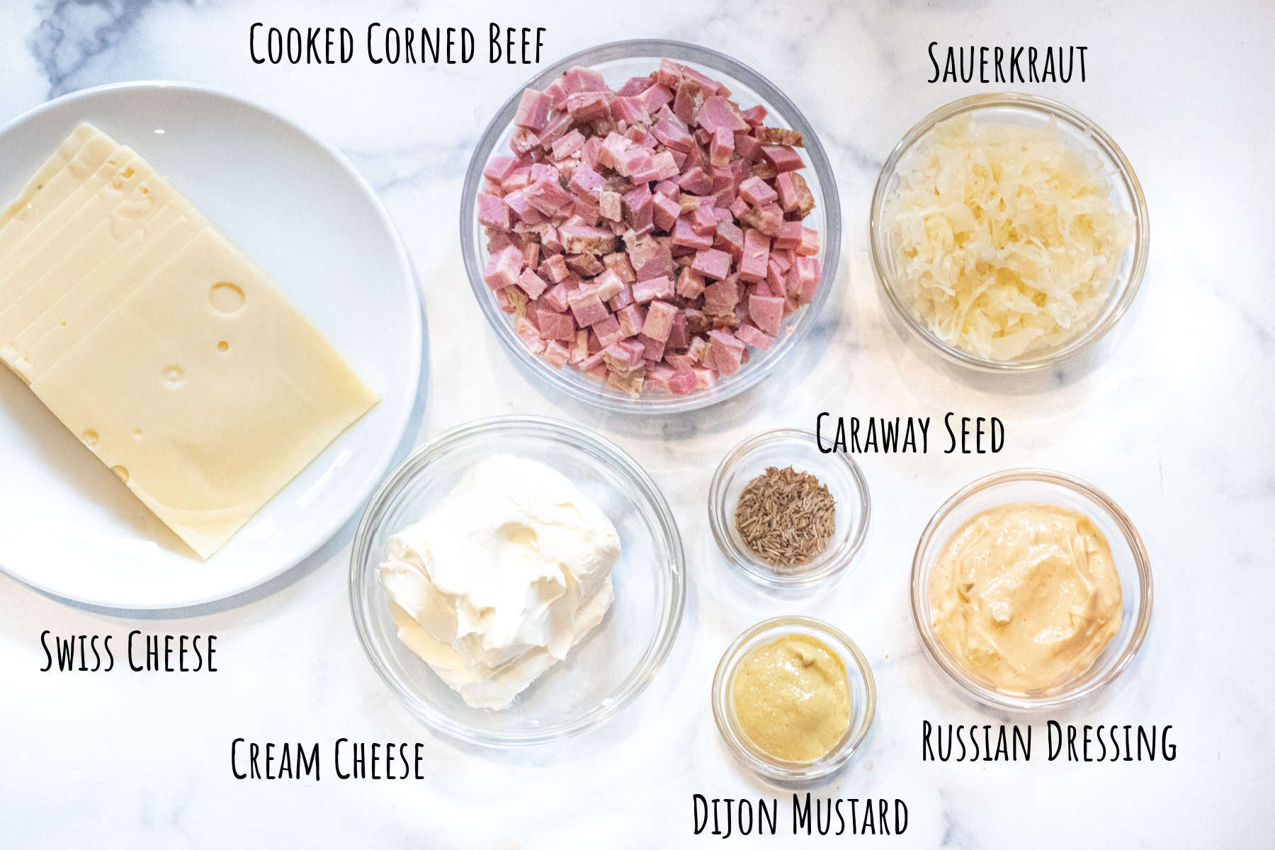 a plate of sliced swiss cheese, a bowl of diced corned beef, sauerkraut, dijon mustard, Russian dressing, cream cheese and caraway seeds.