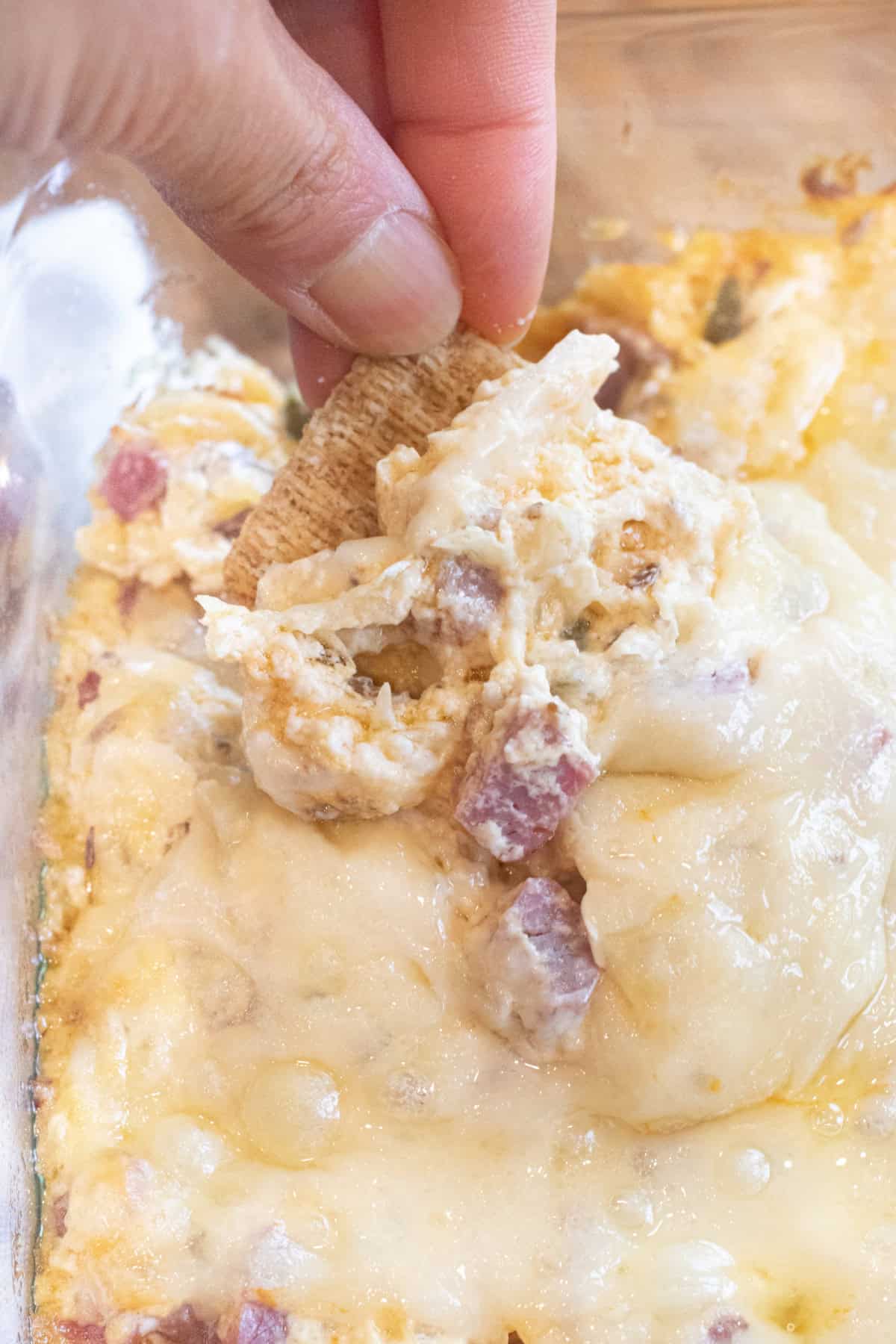 a hand holding a cracker dipping it into a Reuben dip with melted cheese.