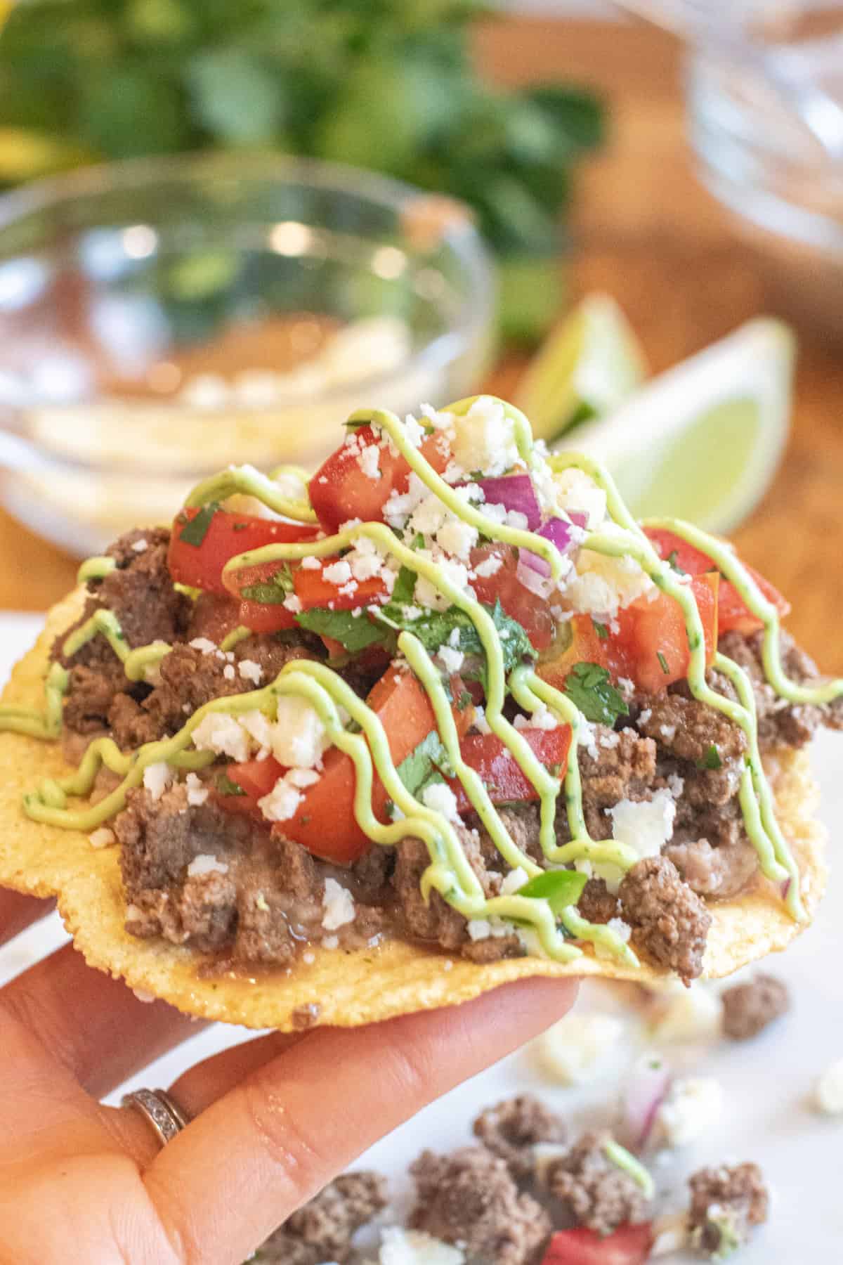 a hand holding a loaded tostada with beef, pico, cotija, avocado cream sauce, and cilantro.