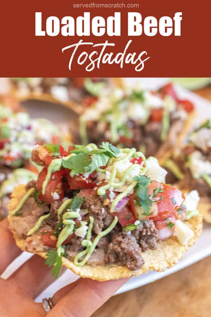 a hand holding a loaded tostada with beef, pico, cotija, avocado cream sauce, and cilantro and Pinterest pin text.