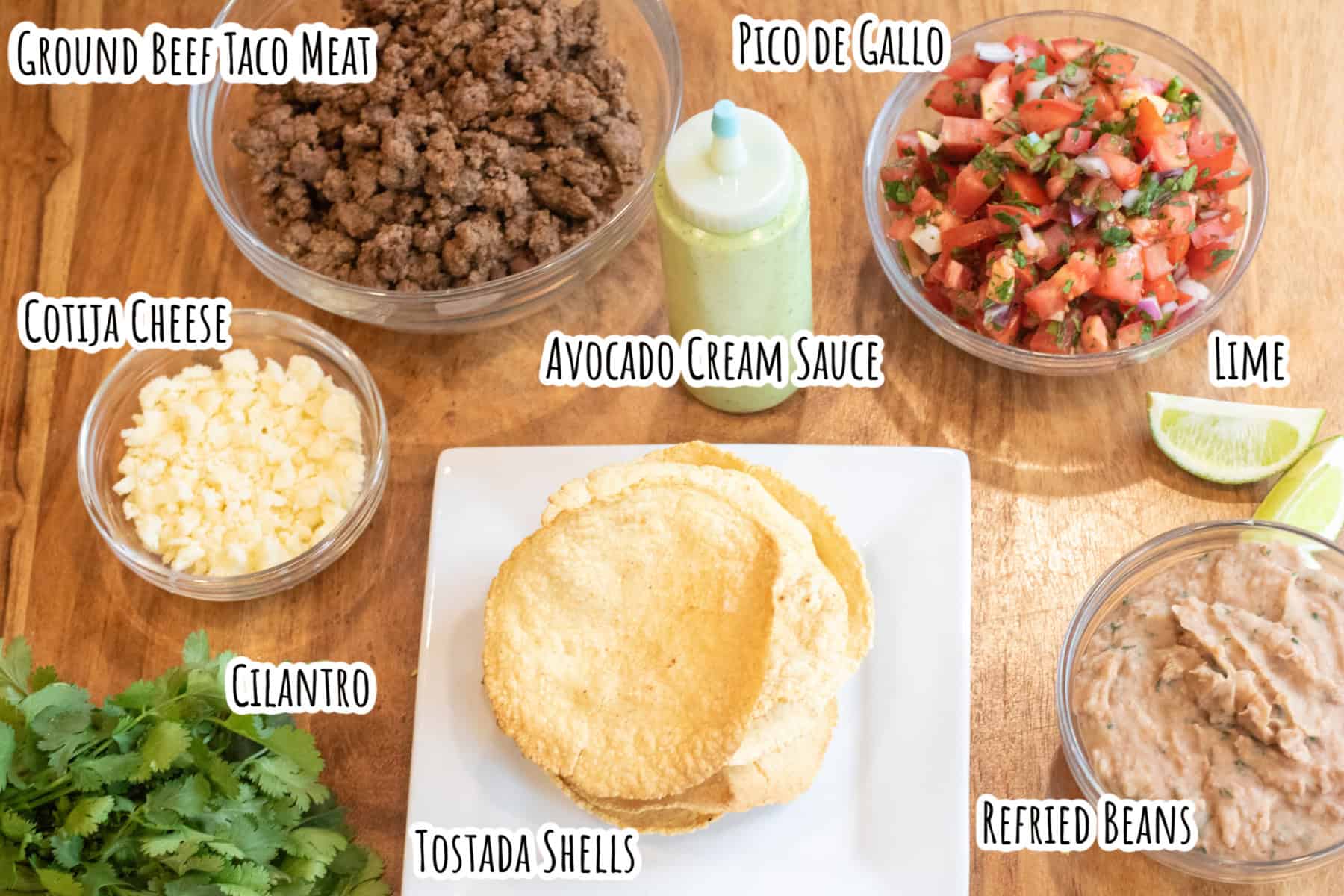 bowls of ground beef, pico de gallo, cotija cheese, refried beans, a plate of tostadas, cilantro, and lime juice and a squeeze bottle of avocado cream sauce all labeled.