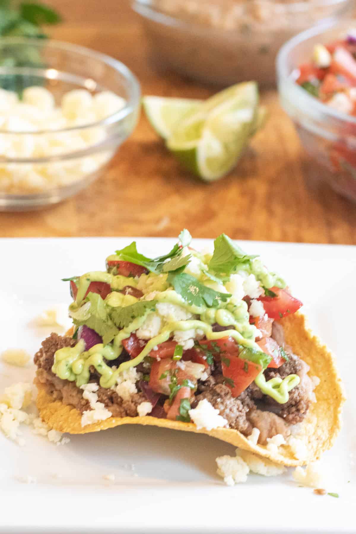 a tostada loaded with ground beef, pico, cheese, and an avocado cream sauce.