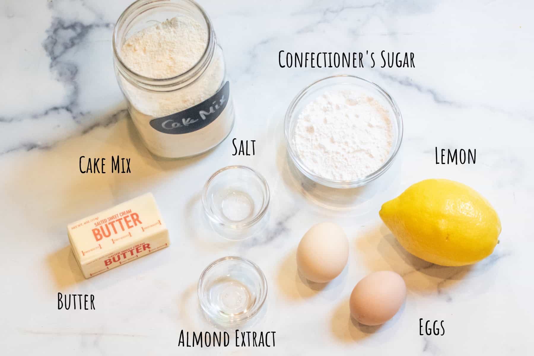 a mason jar of cake mix, bowls of confectioner's sugar, salt, and almond extract, and butter, eggs and lemon all labeled.